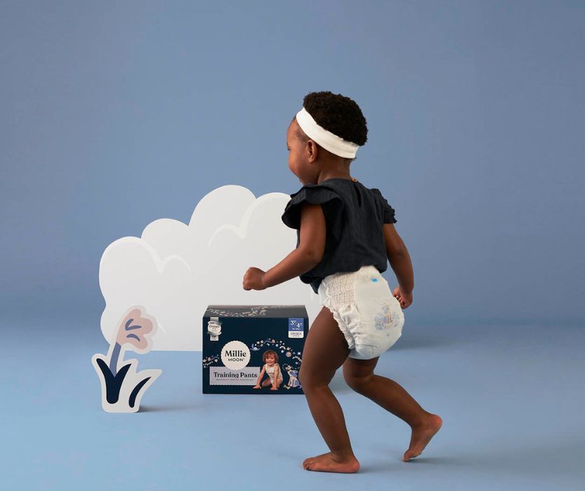 Toddler running through room wearing shirt and training pants in front of cloud and box of diapers.