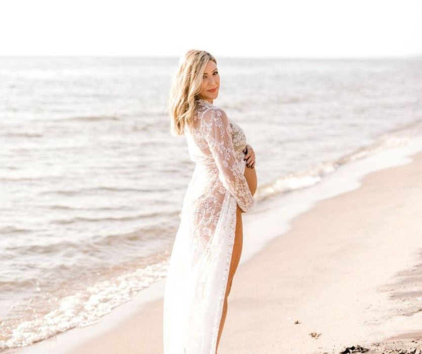 woman holding her baby bump next to the ocean in a lace dress