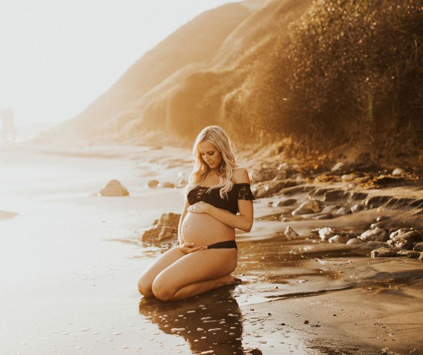 woman kneeling on the beach in a black binkini holding her baby bump