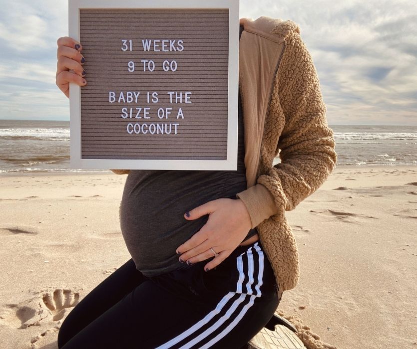 woman on beach holding a sign reading 31 weeks 9 weeks to go baby is the size of a coconut