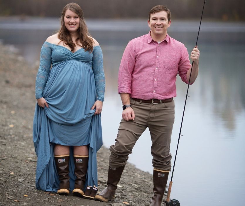 pregnant woman in rubber boots next to man with fly fishing rod beside lake