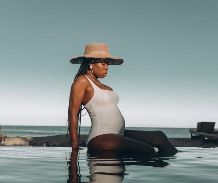 pregnant woman with a straw hat and white bathing suit sitting in an infinity pool by the ocean