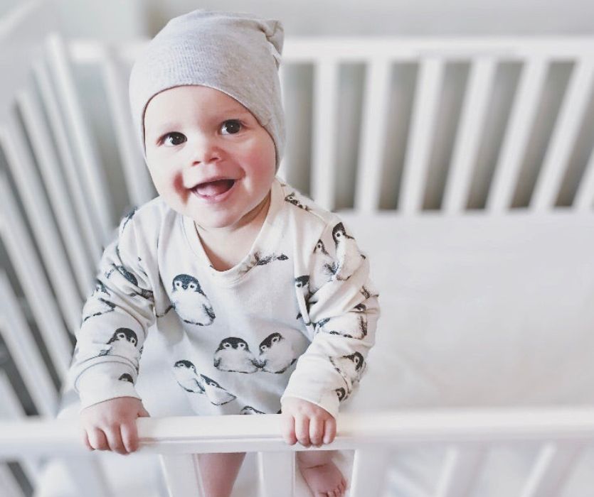 smiling baby stading in a crib wearing a hat and penguin pajamas