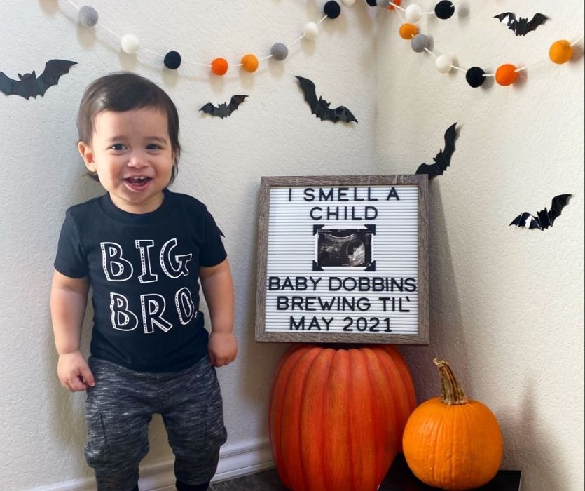 toddler with big bro shirt next to baby announcement sign and pumpkins