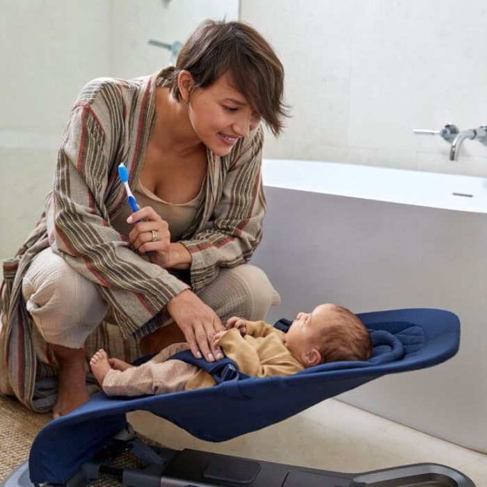 mom with toothbrush looks at baby on bouncer in bathroom