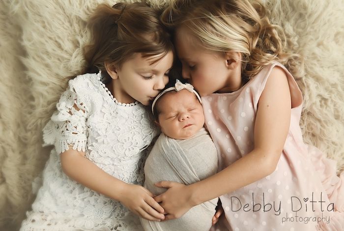 Two sisters with swaddled newborn baby sister posed on furry carpet