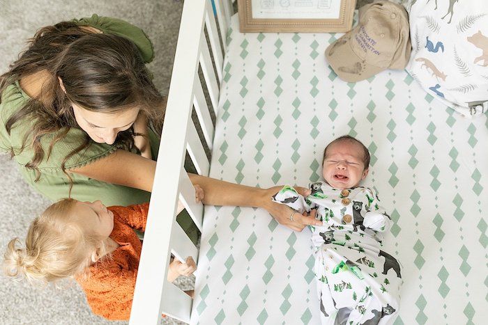 Mom reaching through crib with toddler girl showing her new baby brother