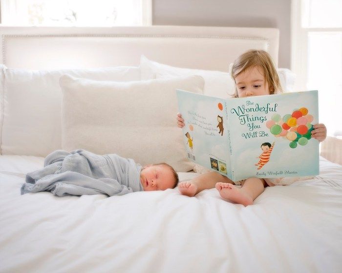 Toddler big sister with large book reading to newborn baby sibling