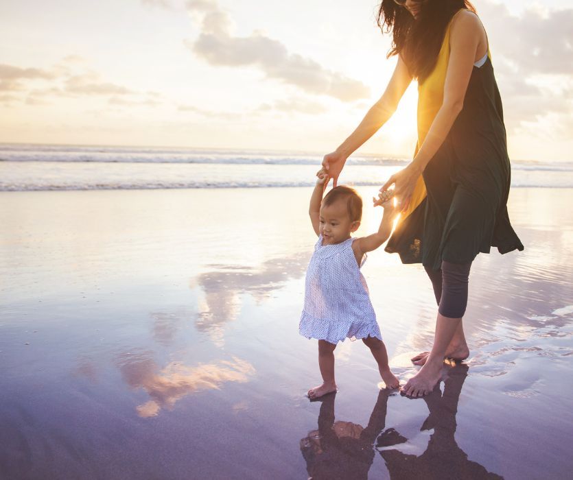 Mom holding the hands of her baby while walking on the beach during vacation.