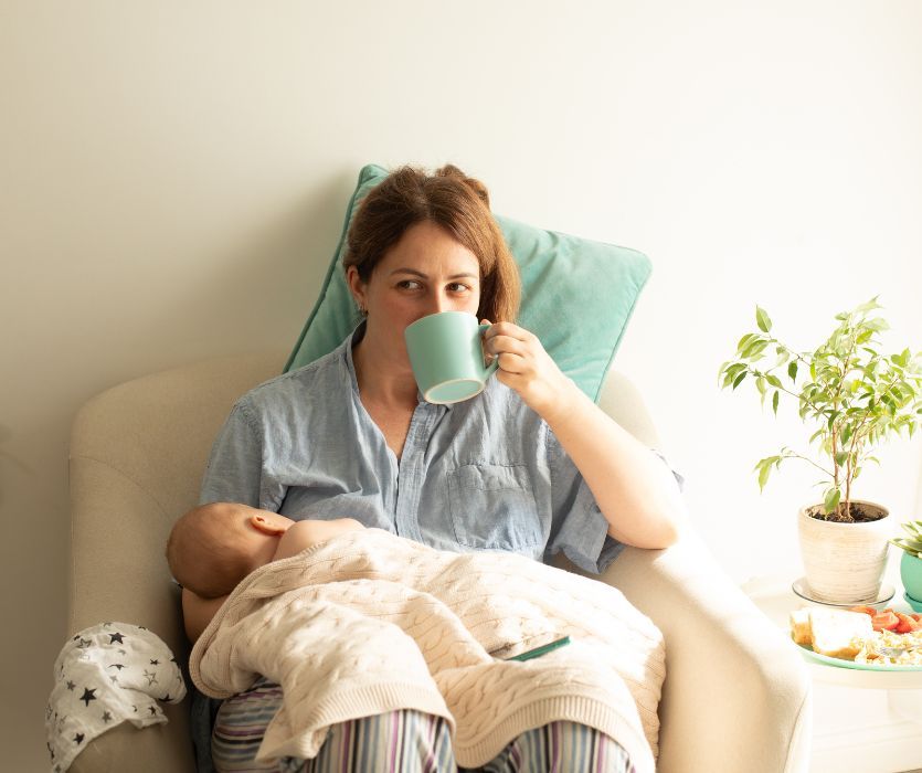 new mom sitting in a chair breastfeeding her newborn baby while drinking coffee