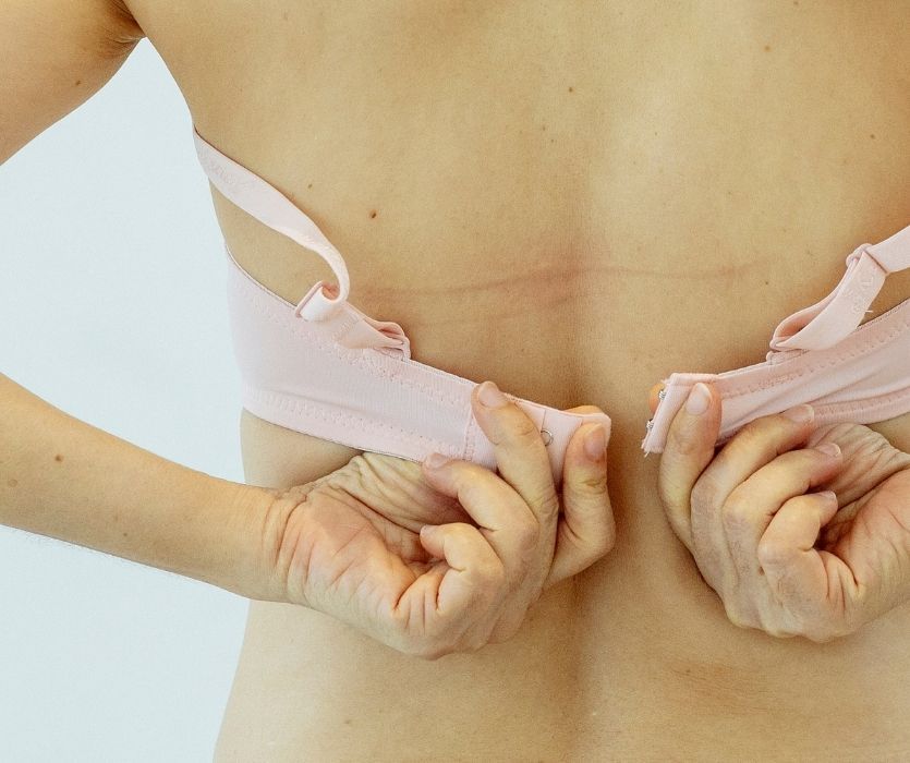 Close-up of a person's back as they reach behind to fasten the clasp of a light pink bra.