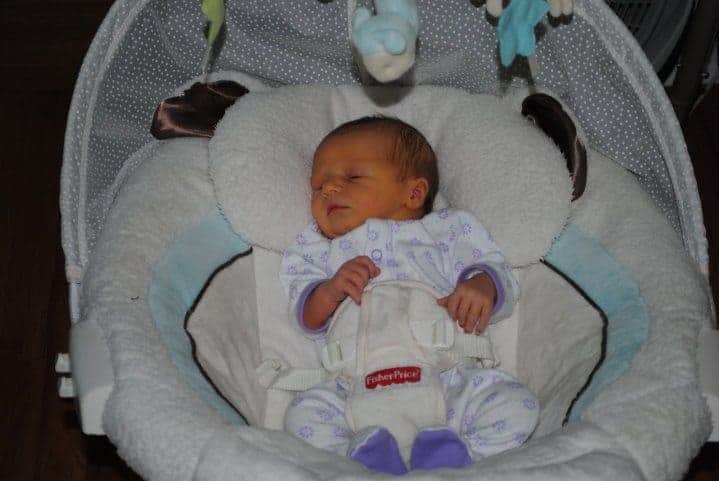 Newborn baby sleeping in a plush Fisher-Price bouncer with hanging toys above.