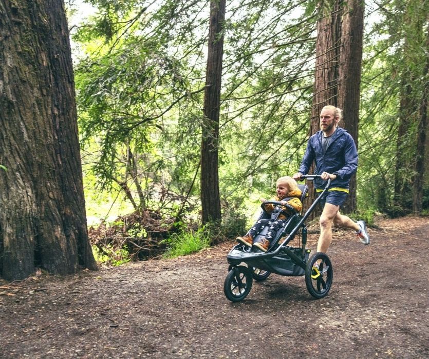 A man running on a wooded trail while pushing a toddler in a black jogging stroller.