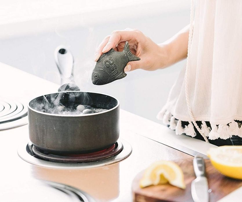 A hand holding a Lucky Iron Fish over a steaming pot of boiling water on a stovetop.