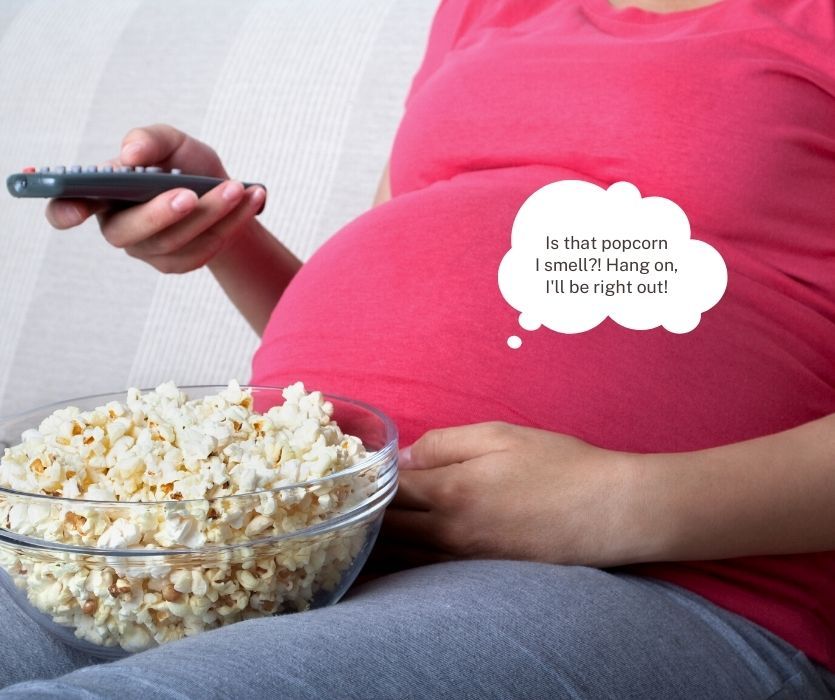 Pregnant person holding a TV remote and popcorn. A thought bubble says: Is that popcorn I smell?! Hang on, I'll be right out!