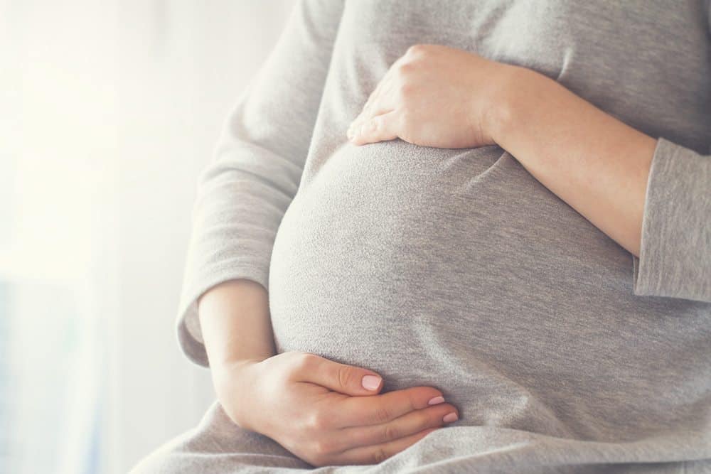 A person in a gray dress cradling a large third-trimester pregnant belly with both hands.