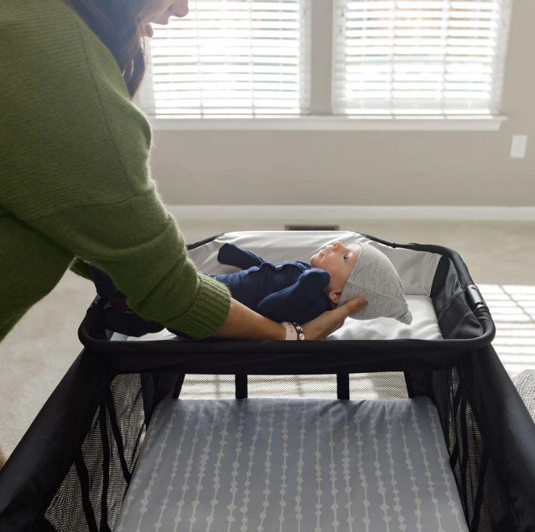 A woman places a reclining infant into the bassinet attachment of a 4moms Breeze Plus portable playard.