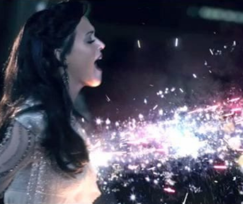 Katy Perry singing with her mouth open as bright, colorful sparks fly toward a dark night sky.