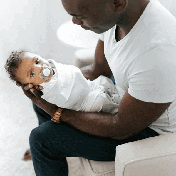 A man sits in a chair holding a swaddled infant with a pacifier in its mouth.