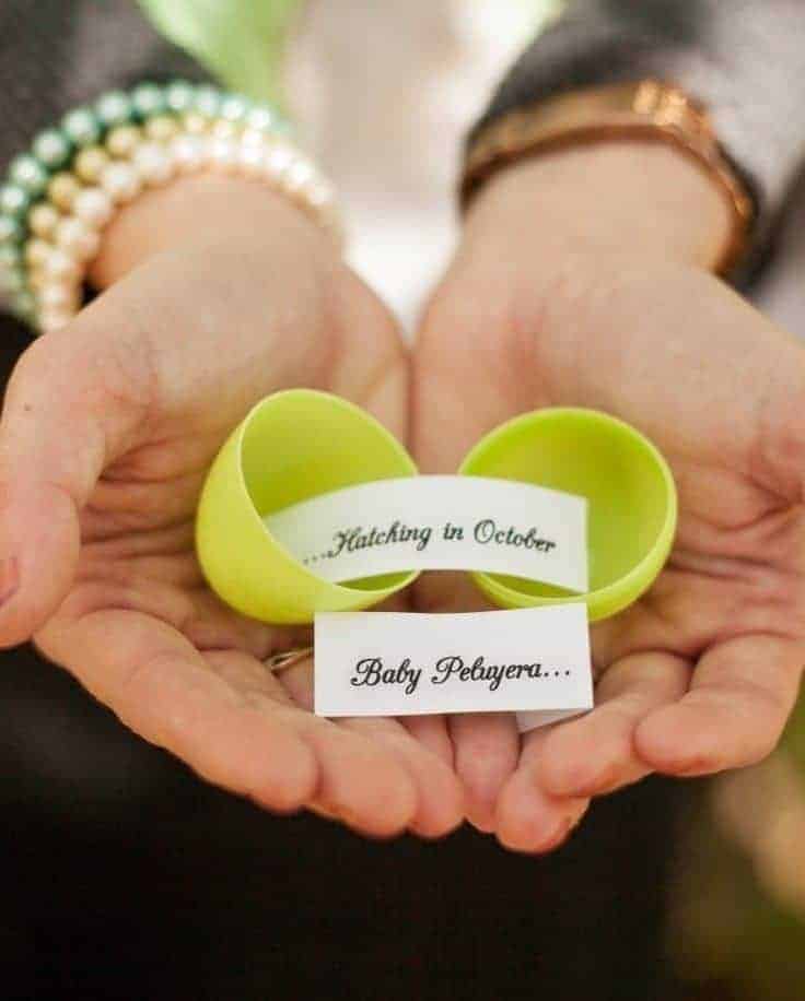 Two hands holding an open green plastic egg with paper strips reading, Hatching in October Baby Peluyera.