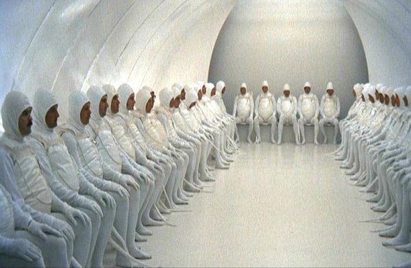 Rows of men dressed in all-white sperm costumes sitting in a curved, white futuristic room.