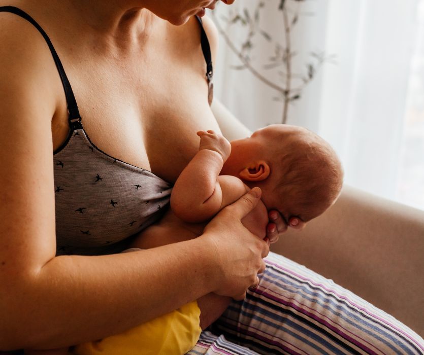 A woman in a nursing bra breastfeeding an infant while sitting in a chair.