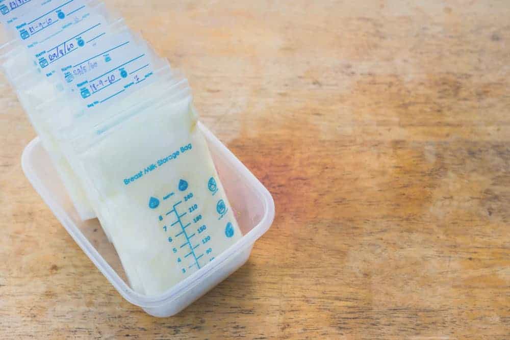 Several labeled breast milk storage bags filled with milk, standing in a small plastic container on a wooden surface.