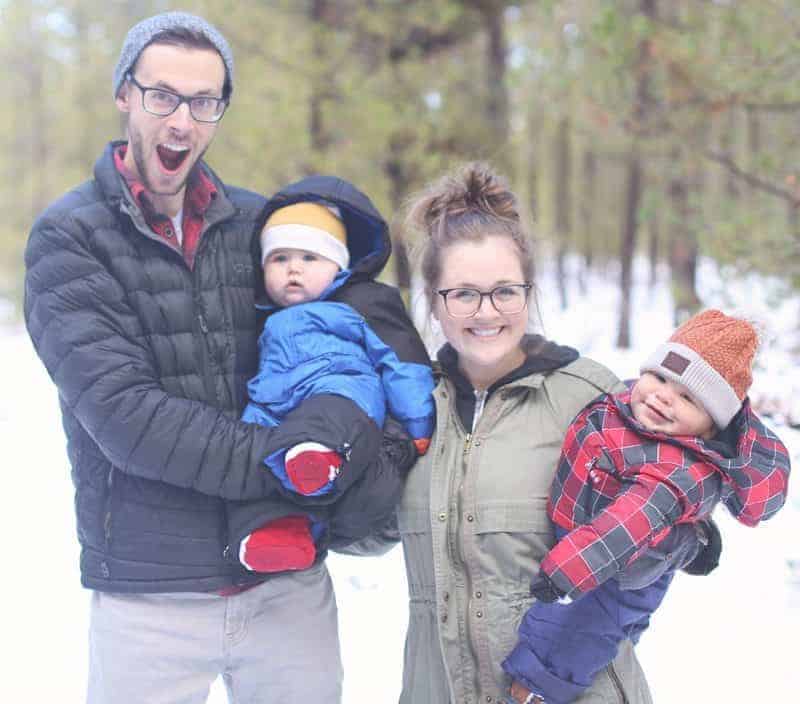 A man and woman smiling in the snow while each holding a baby dressed in winter gear.