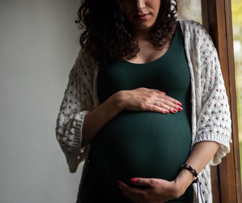 Pregnant woman in a green dress and white cardigan holding her belly.
