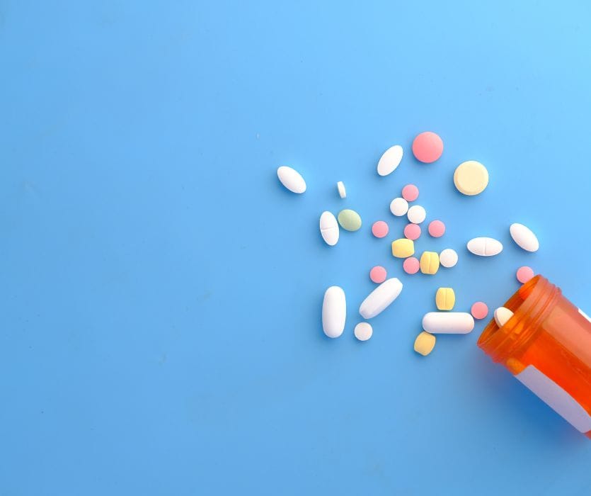 Assorted colorful pills and white capsules scattered next to an open orange prescription bottle on a blue background.