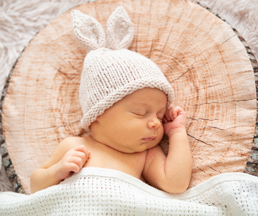 Sleeping newborn wearing a white knit bunny ear hat, lying on a tree ring print pillow under a white blanket.