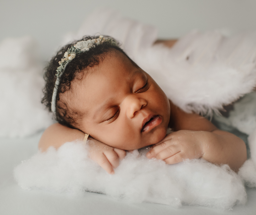 Newborn with dark curly hair wearing a floral headband, sleeping on white fluffy material resembling clouds.