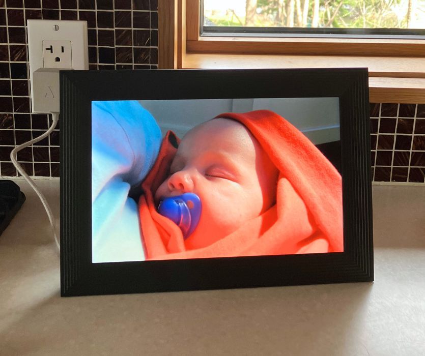 Aura digital photo frame on a kitchen counter displaying a photo of a sleeping baby wrapped in an orange blanket.