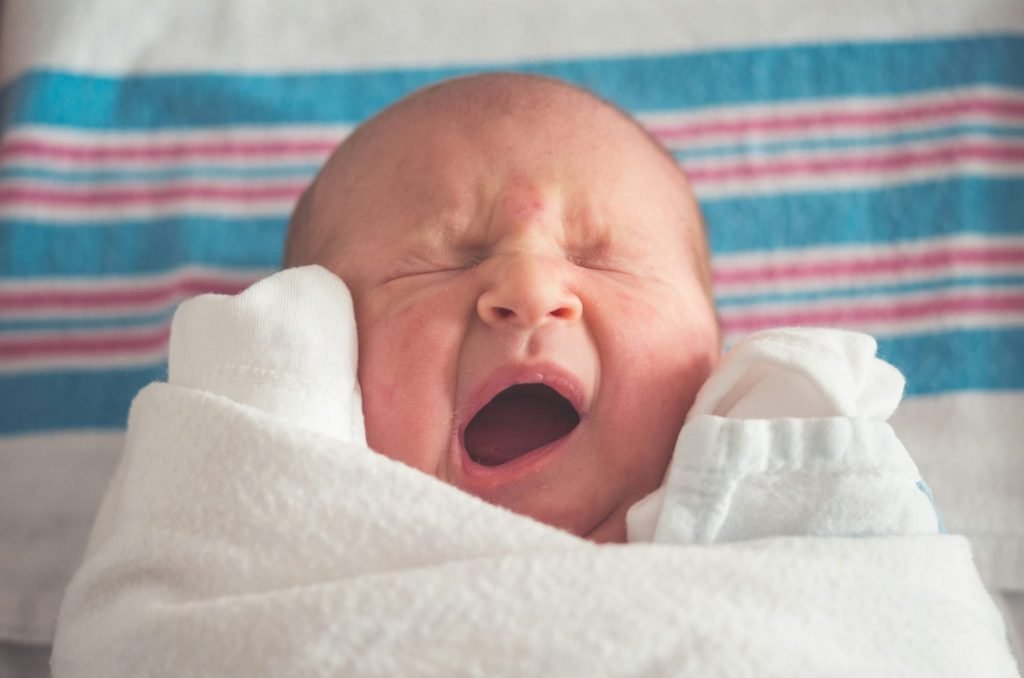 Swaddled newborn baby yawning with eyes closed, lying on a blue and pink striped hospital blanket.