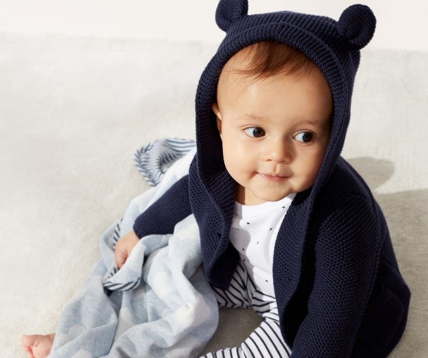 Baby wearing a navy garter-stitch cardigan with bear ears on the hood, sitting on a rug with a blue patterned blanket.