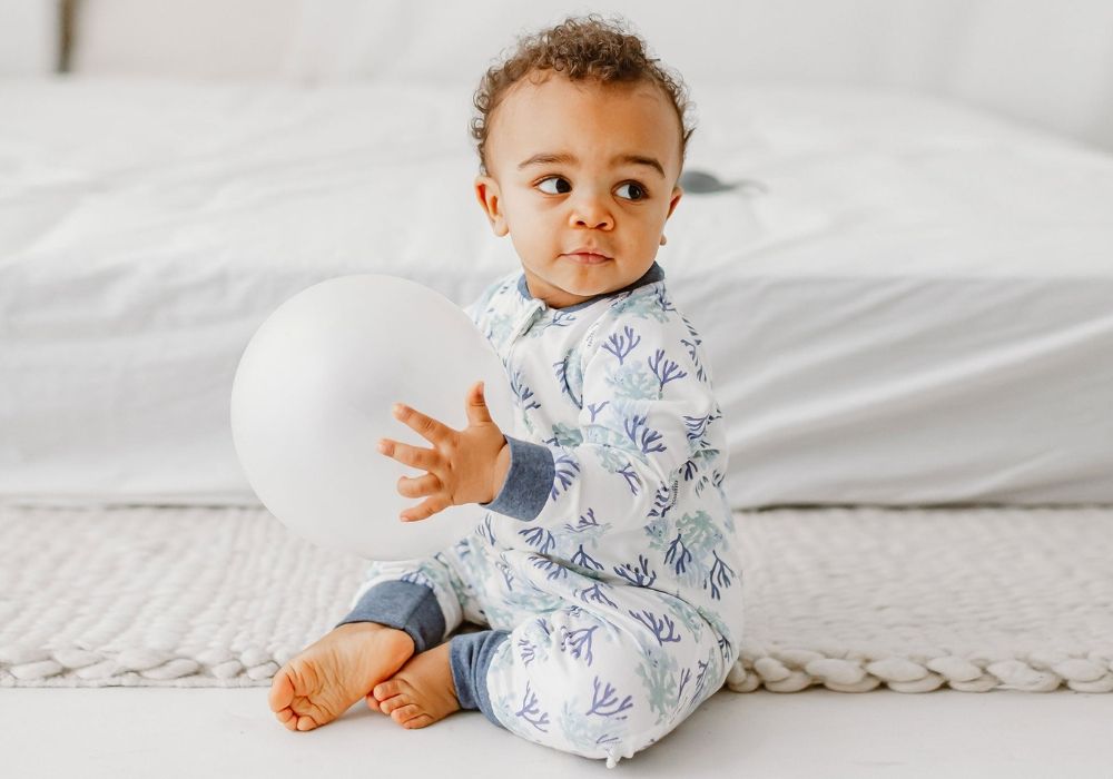 Toddler in a patterned bamboo romper sitting on a rug and holding a white balloon.
