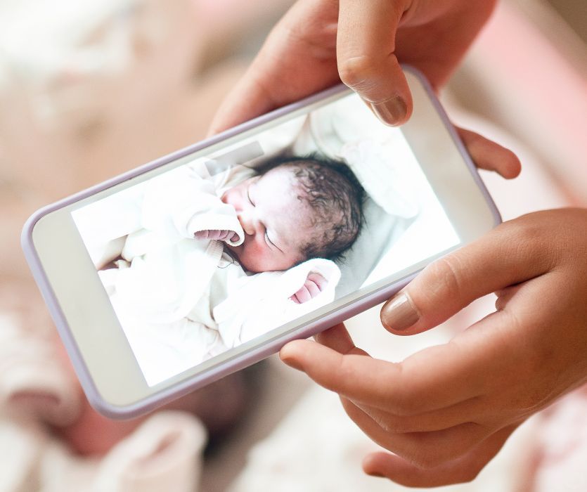 Hands holding a smartphone displaying a photo of a sleeping newborn baby in white clothing.