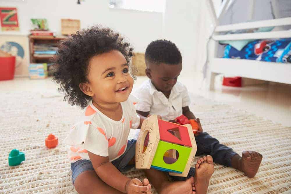 Two young children playing with a wooden shape sorter toy on a rug.