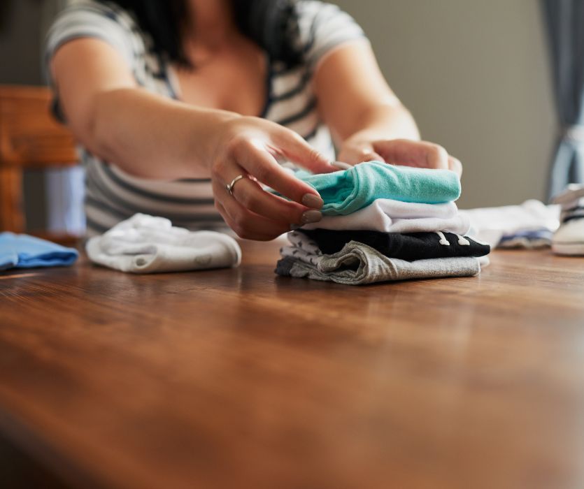 A woman sitting at a wooden table folding a stack of colorful baby clothes.