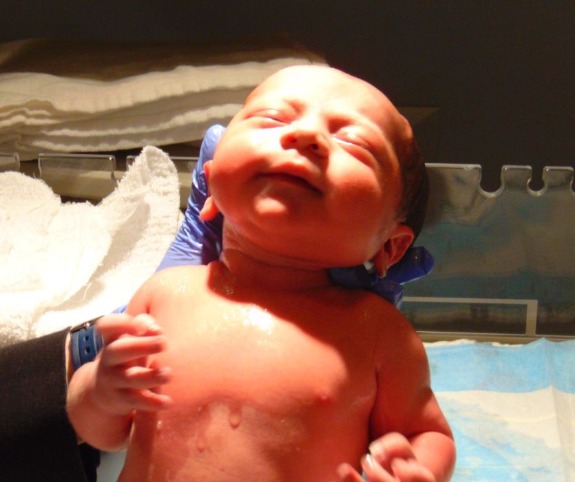 A newborn son lies in a hospital bassinet while a gloved hand supports his head, his face scrunched in the bright light after birth.