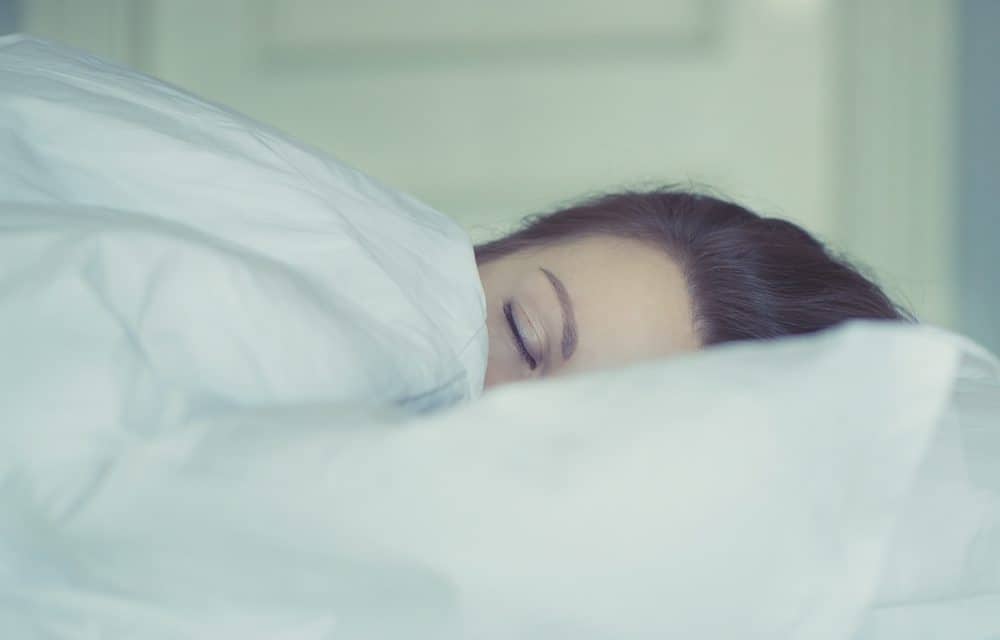 A person with dark hair sleeps in a bed with their face partially covered by white blankets.