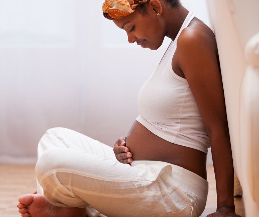 Pregnant woman sitting on the floor leaning against a wall, looking down and holding her belly.