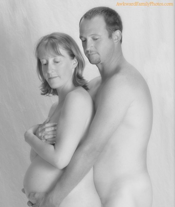 Black and white photo of a shirtless man standing behind and hugging a topless pregnant woman.
