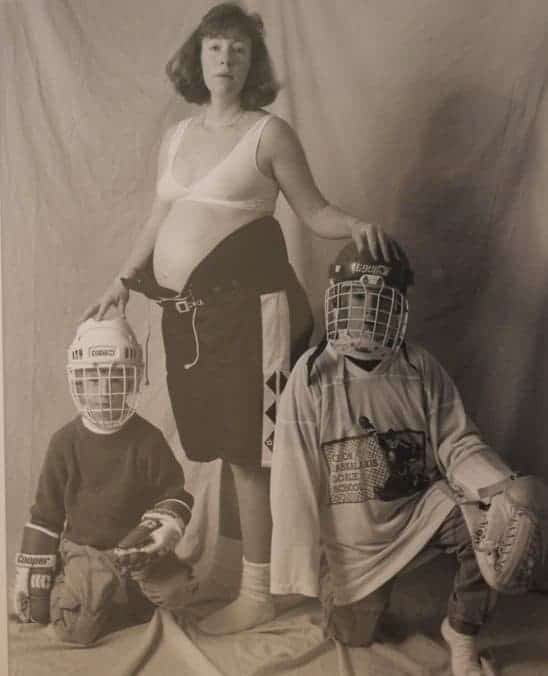 Pregnant woman in a bra and hockey shorts standing with two children wearing hockey gear and helmets.