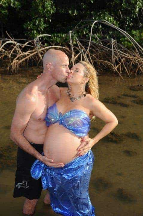 Shirtless man and pregnant woman in blue mermaid costume kissing while standing in water by mangrove roots.
