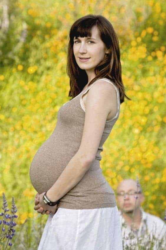 Pregnant woman in profile in a field of yellow flowers, while a man's head peeks out near her waist in the background.