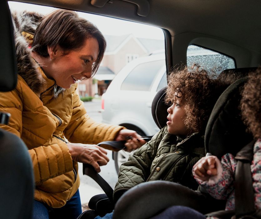 A woman buckling a child wearing a thick winter coat into a car seat, illustrating a safety mistake.