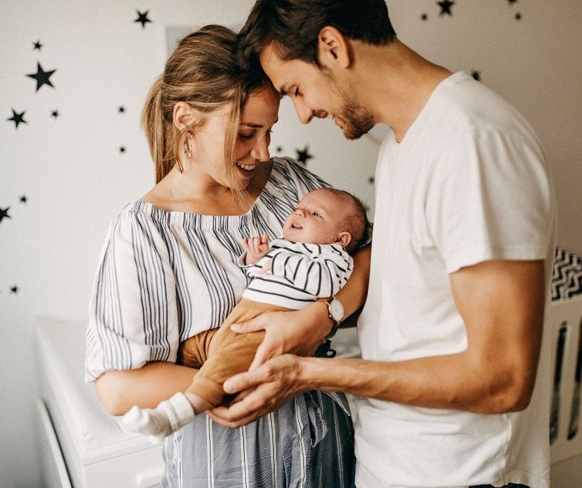 Smiling man and woman looking down at a newborn baby they are holding in a nursery.