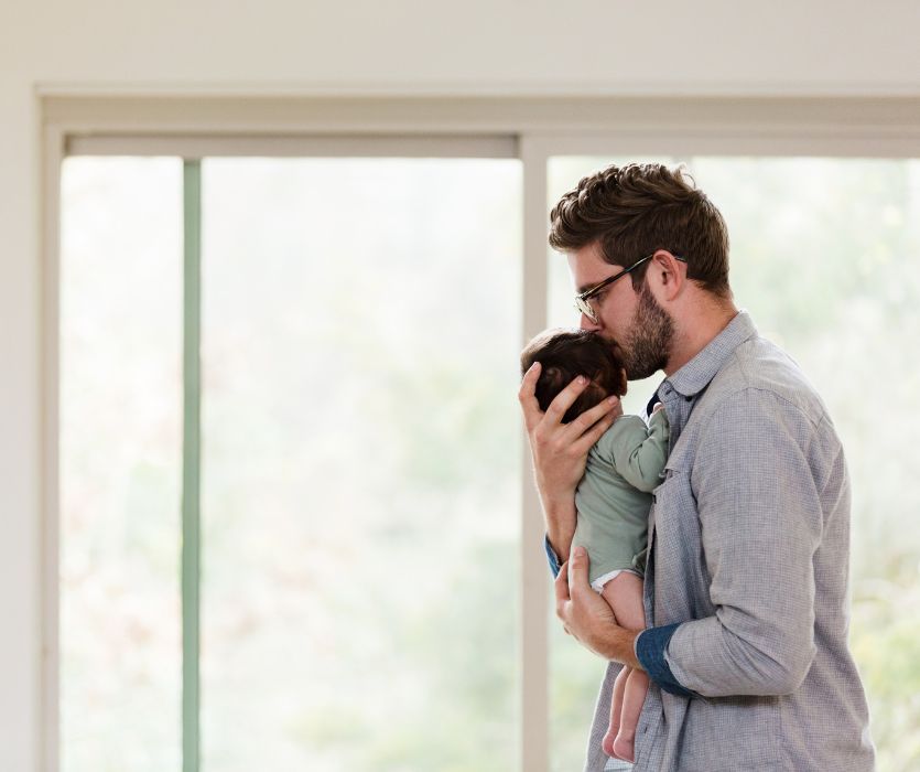 A father wearing glasses kisses the head of his newborn baby while holding them in front of a window.