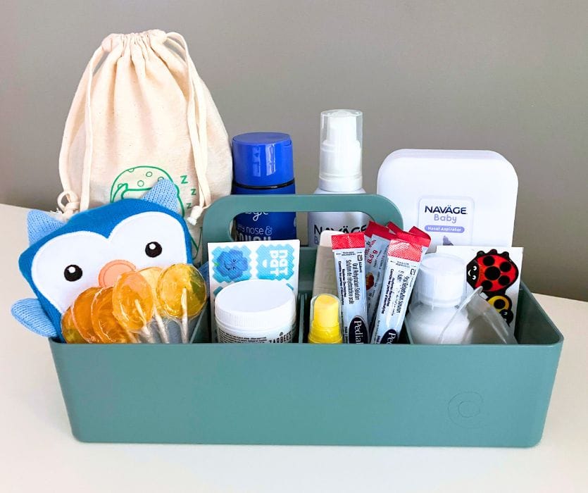 A green plastic caddy filled with lollipops, nasal spray, electrolyte packets, and other pediatric sick care supplies.